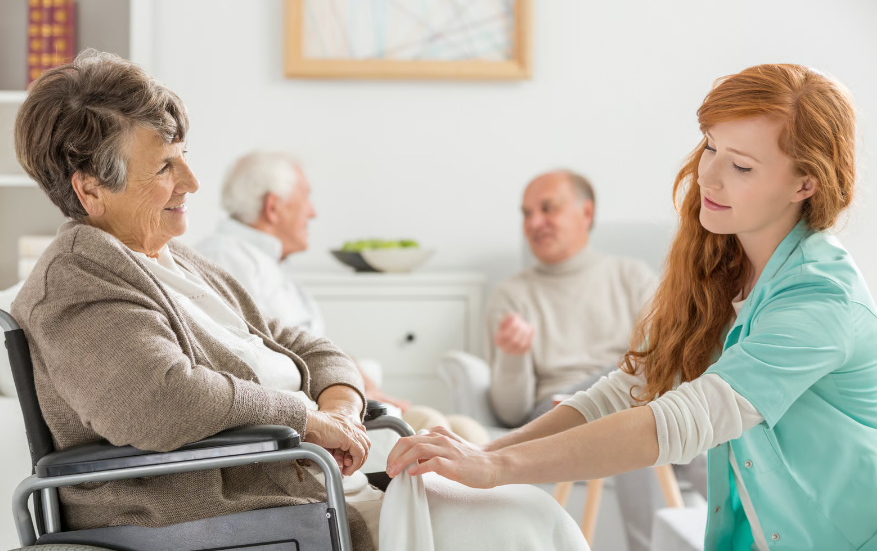 A friendly hospice nurse in green scrubs tucking a blanket around a smiling senior woman in a wheelchair.