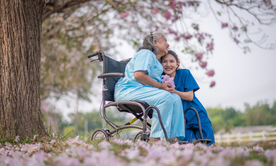 A young female nurse in blue scrubs kneeling beside a senior woman in a wheelchair under a blossoming tree, illustrating the emotional and physical support of palliative and hospice care.
