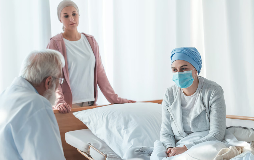 A senior doctor consulting with a young female patient in a headscarf and mask while a caregiver stands supportively by the bedside.