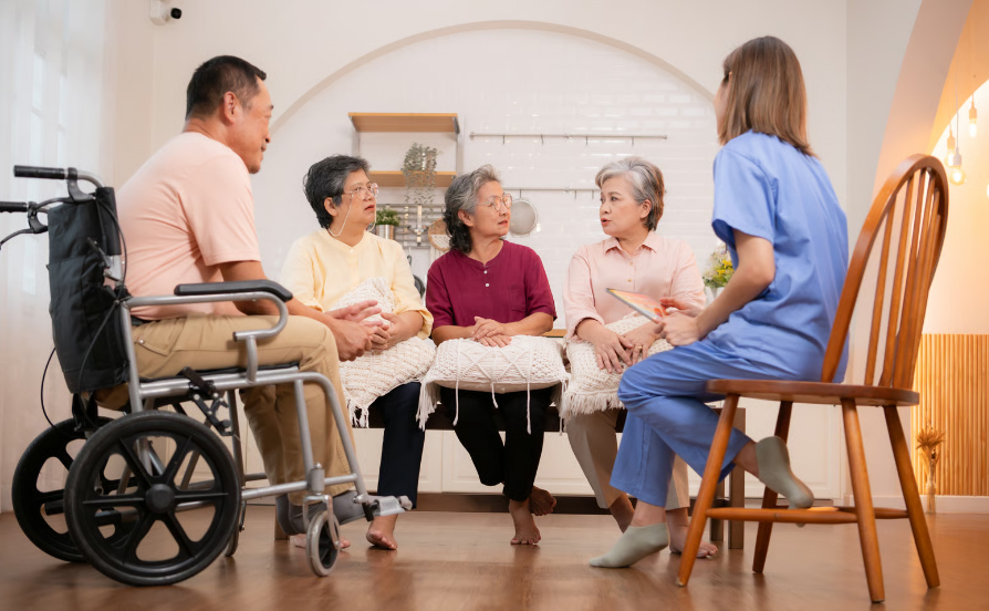 A group of elderly Asian residents, including a man in a wheelchair and three women, sitting in a living room having a group discussion with a female nurse in blue scrubs holding a tablet.