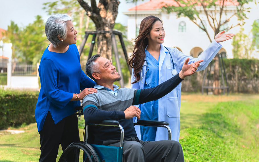 An elderly Asian man in a wheelchair smiling and pointing forward alongside his wife and a female doctor in a park.