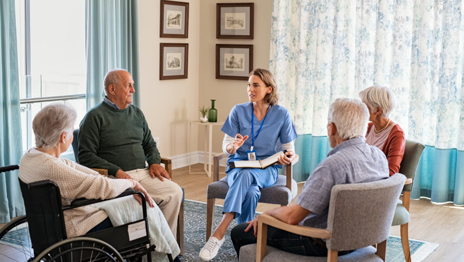 A female nurse in blue scrubs holds a binder while leading a group discussion with senior citizens seated in a circle at an assisted living facility.