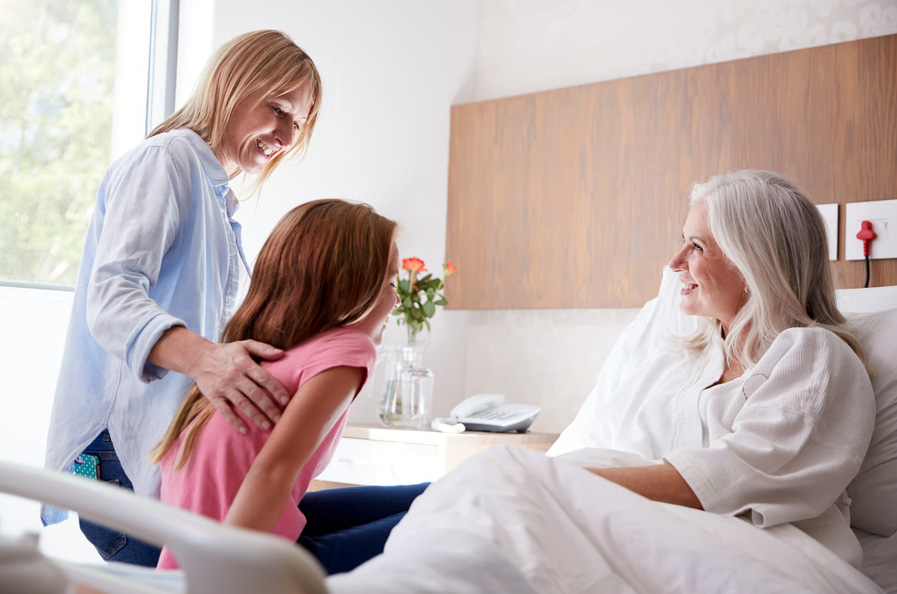 A mother guiding her young daughter as they visit a smiling grandmother resting in a hospice bed.