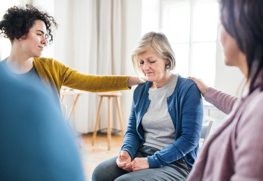 Two women placing comforting hands on the shoulders of an emotional senior woman during a hospice caregiver support session.