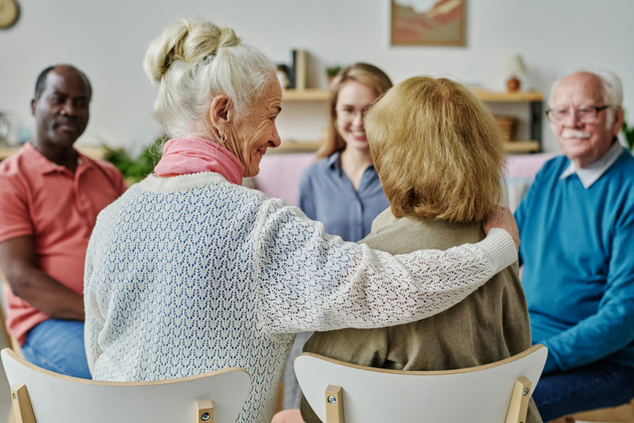 An elderly woman gently placing a hand on another senior's shoulder during a hospice chaplain emotional support session in a circle.