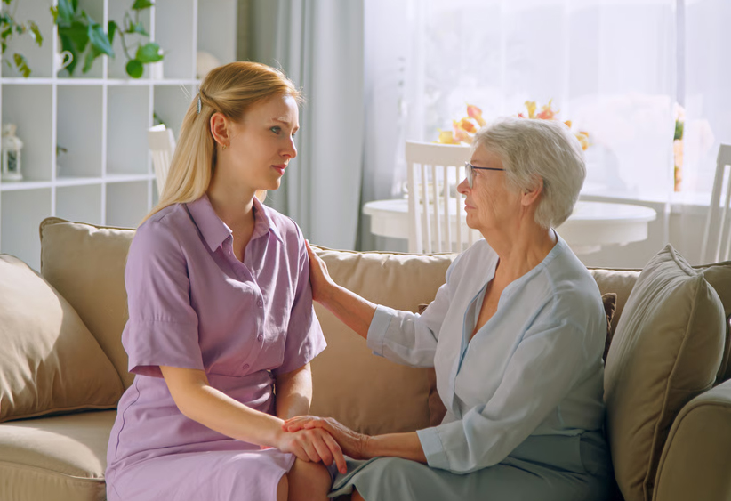 Younger woman offering supportive care and holding hands with an elderly woman on a couch in a home setting.