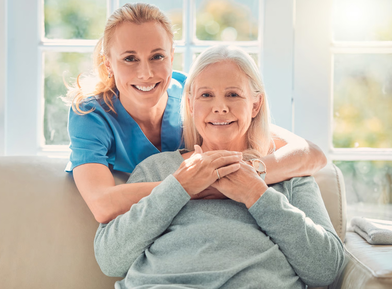A smiling female hospice nurse in blue scrubs hugging a senior female patient sitting comfortably on a couch.