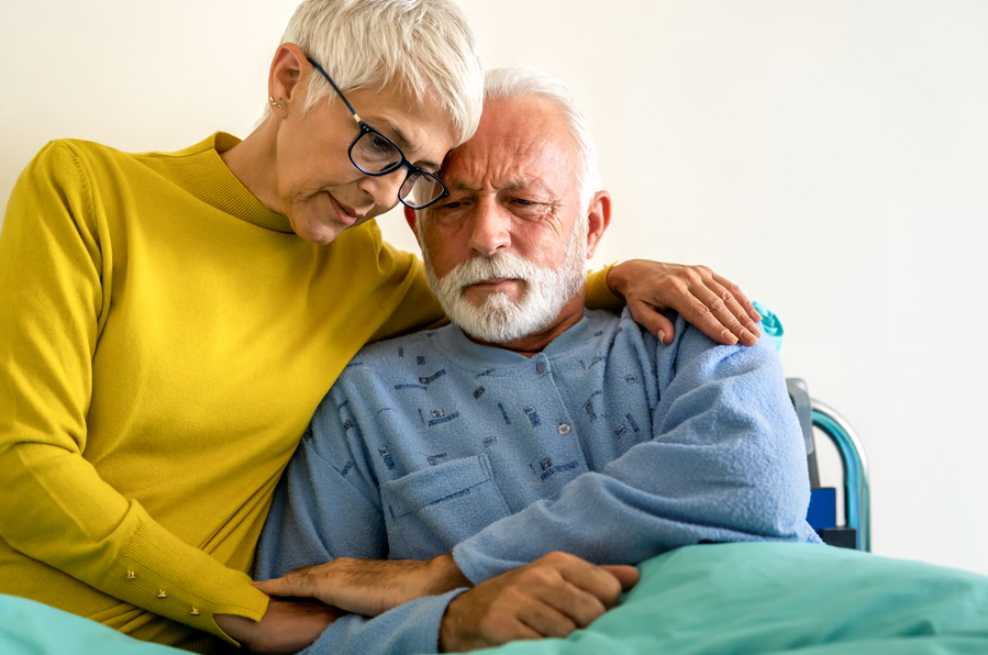 An elderly woman with glasses placing her hand around a somber senior man in a blue hospital gown to comfort him.