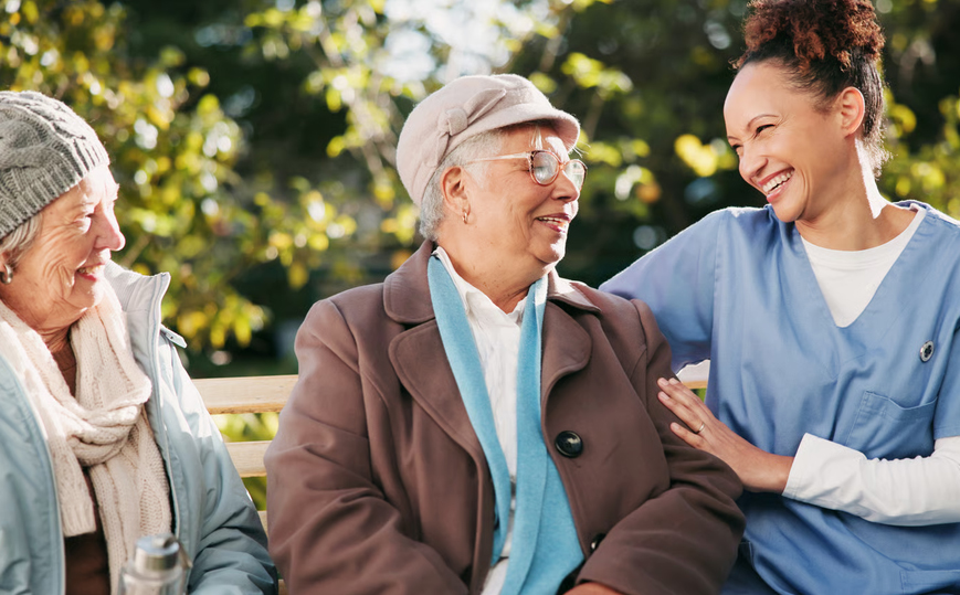 A friendly hospice nurse in blue scrubs smiling and resting her hand on a senior woman sitting outdoors with family