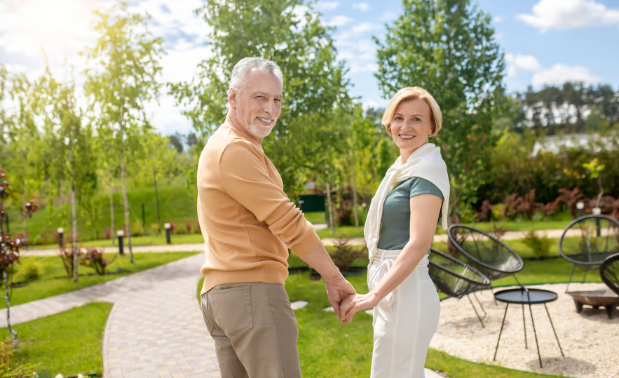 Senior couple smiling and holding hands while walking outdoors along a paved garden path, representing peace of mind with hospice support.